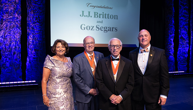 Chair Wilkerson, Goz Segars, JJ Britton and President Clements all wearing formal attire stand in front of a projection screen that says 'Congratulations JJ Britton and Goz Segars'.