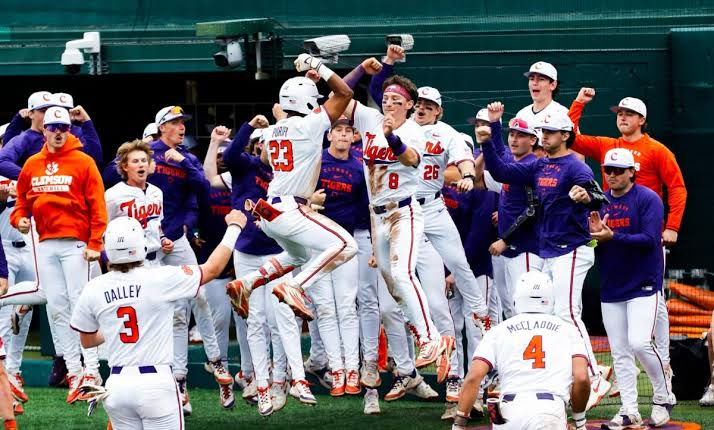 Clemson Tigers baseball players and coaches celebrate in the dugout, jumping and raising their arms after a big play.