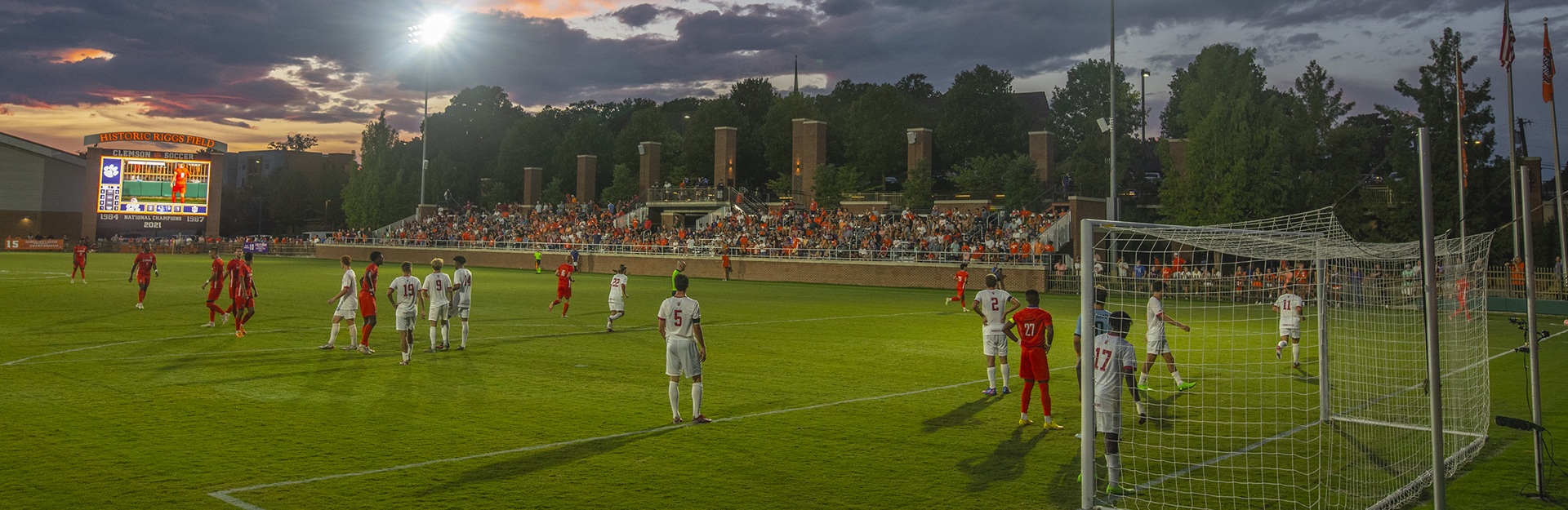 Soccer players on a field during a match at Historic Riggs Field with spectators in the stands at sunset.