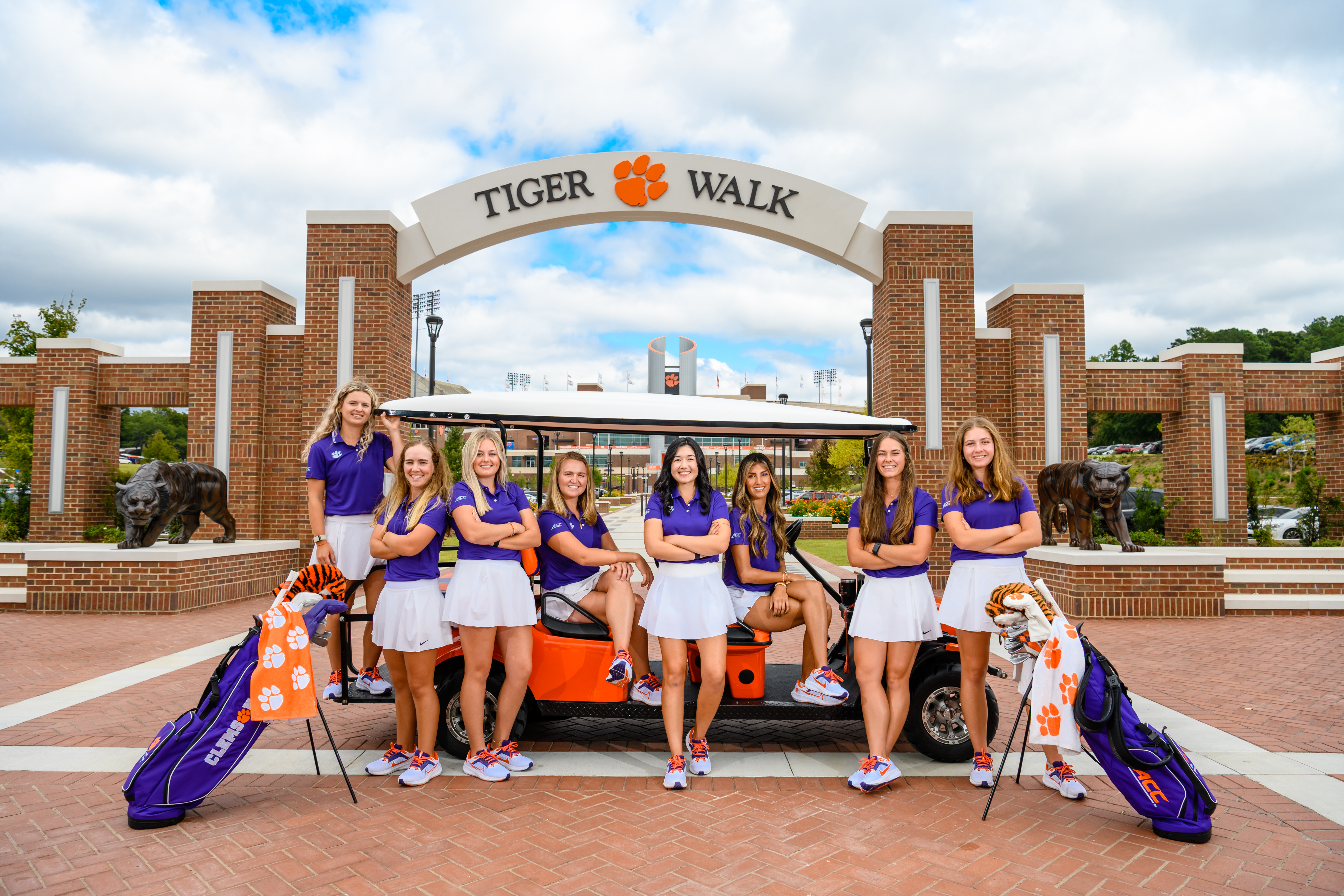 Women's golf team standing on Tiger walk in front of and riding on a golf cart. Two golf bags bookend the group of golfers.