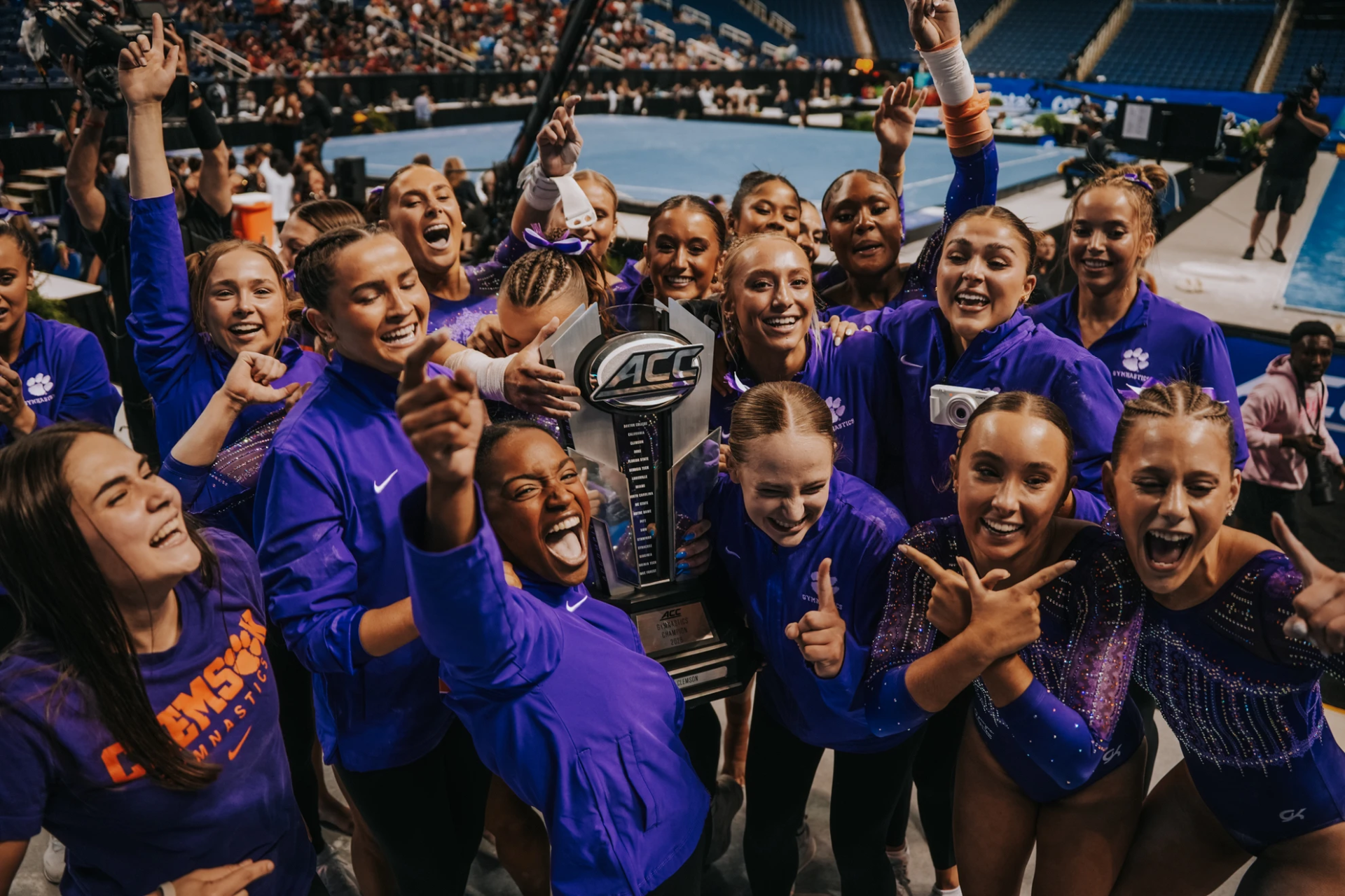 Clemson women's gymnastics team in purple celebrates around an ACC trophy near the competition floor, cheering with arms raised in an arena.