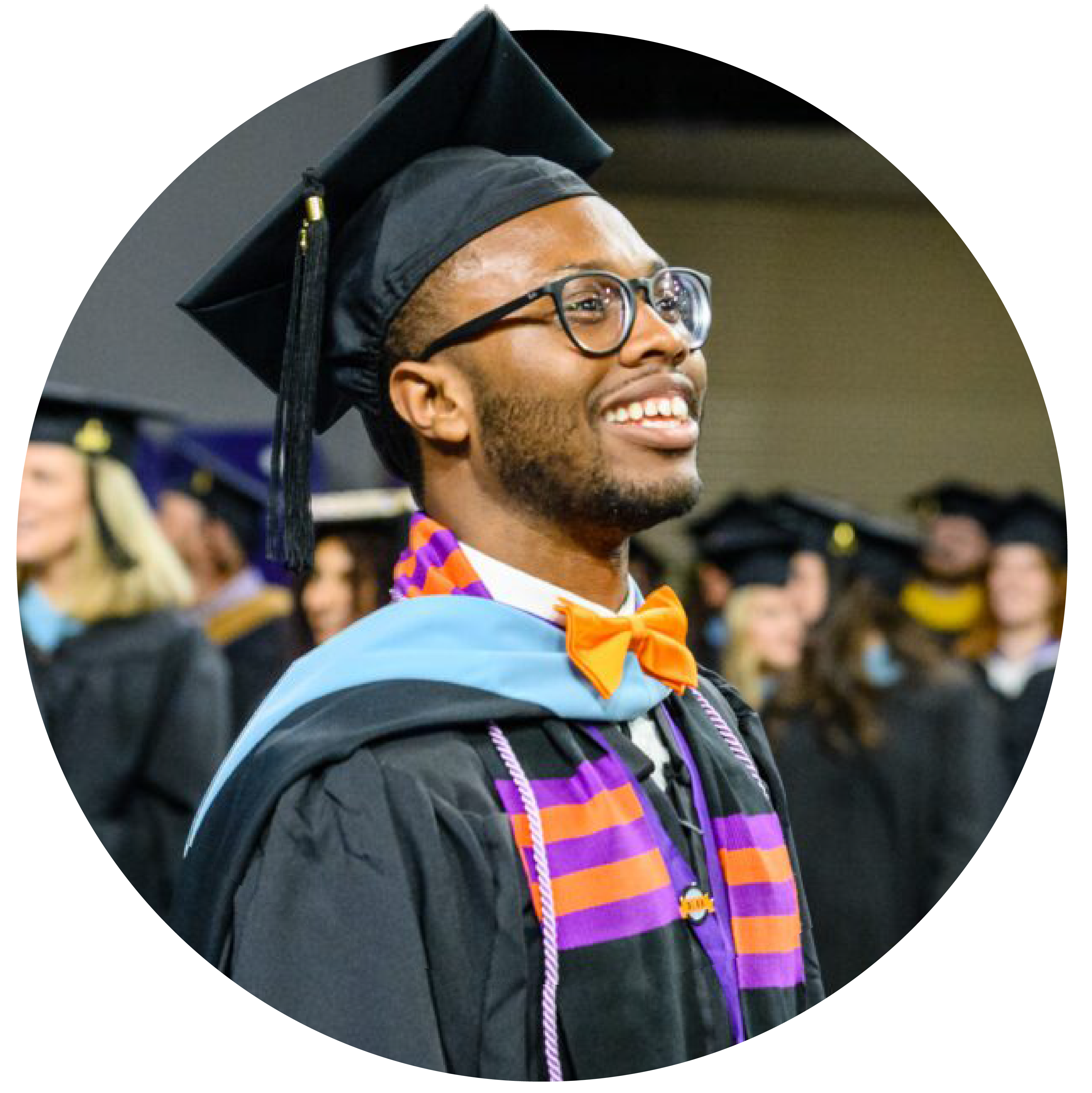 A smiling male student wearing glasses, a black graduation cap and gown, and a purple-and-orange stole at a commencement ceremony.