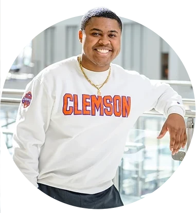 A male student wearing a white sweatshirt with Clemson written in orange letters poses in front of a glass railing in an academic building.