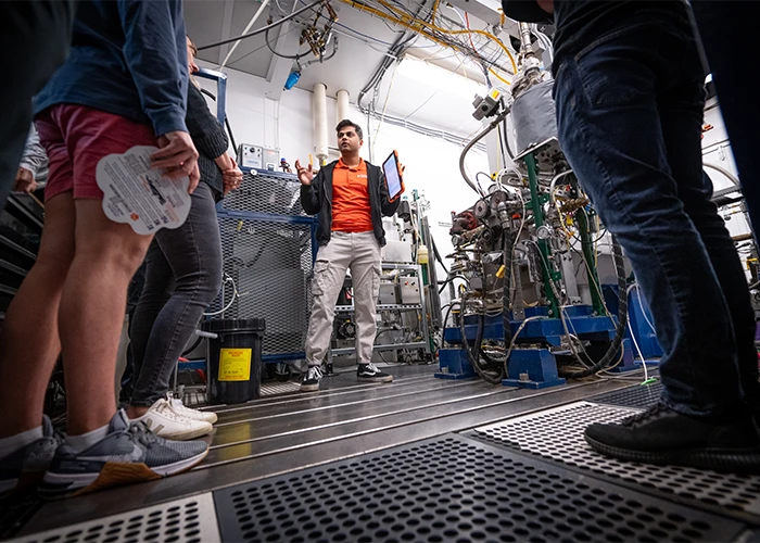 A professor describes a large machine in a warehouse with a group of students' legs in the foreground.