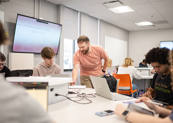 A professor stands beside a student and writes in his notebook, while other students nearby work on computers and tablets.