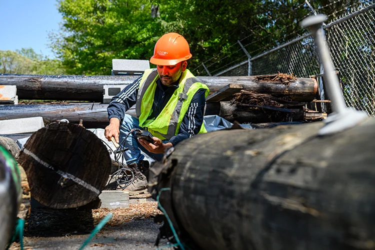 A student in an orange construction helmet and safety vest examines a device mesuring readings from a wooden powerline pole