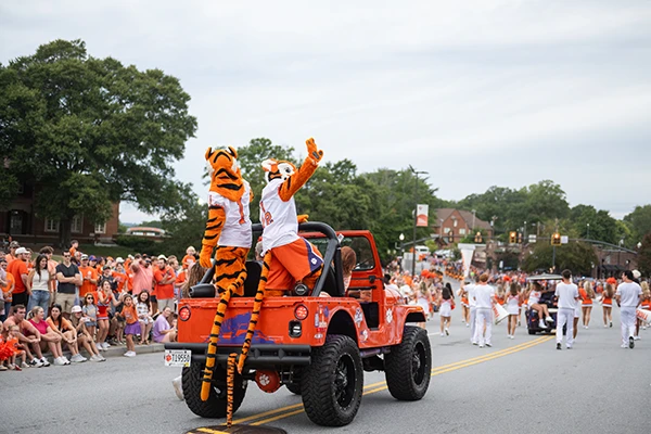 Two Clemson Tiger mascots wave to parade crowds from the back of an orange jeep with the top down.