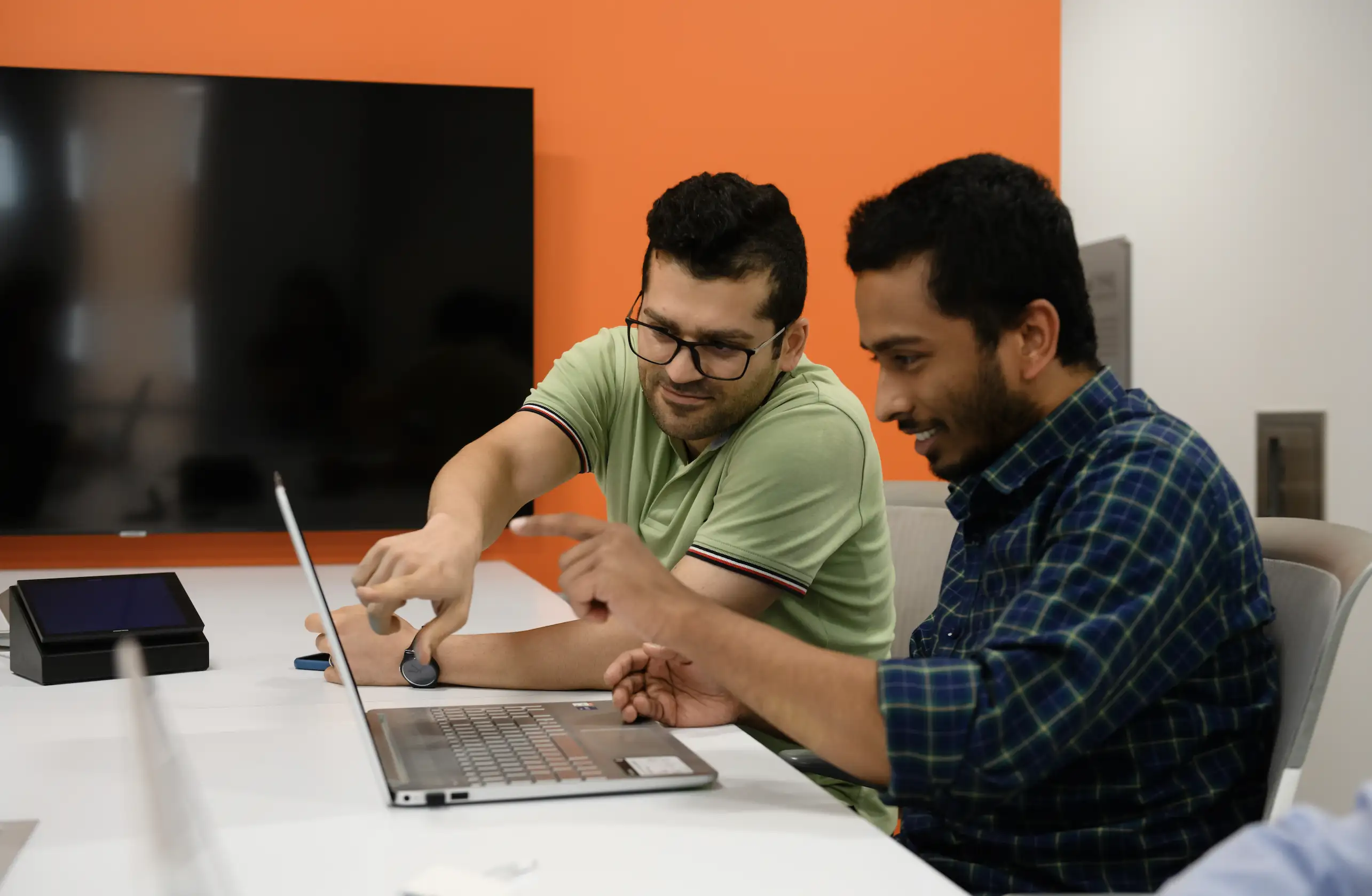 Two men sitting beside each other point and look at the screen of a laptop, with an orange wall and large TV in the background.