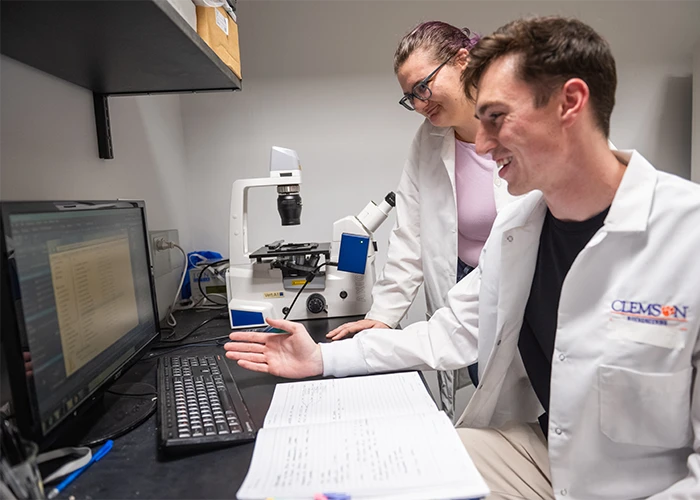 Two graduate students in lab coats use a computer, notebook, and microscope to do research.