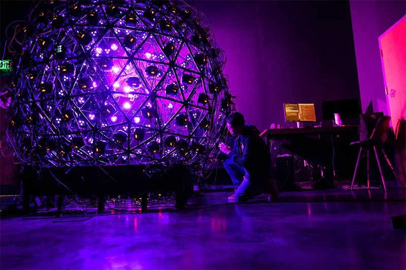 Two shadowed figures kneel to inspect a large orb consisting of purple spotlights and several cameras.