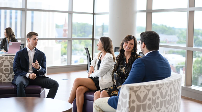 Two pairs of Clemson graduate students converse with each other in a student lounge with floor to ceiling windows overlooking campus.