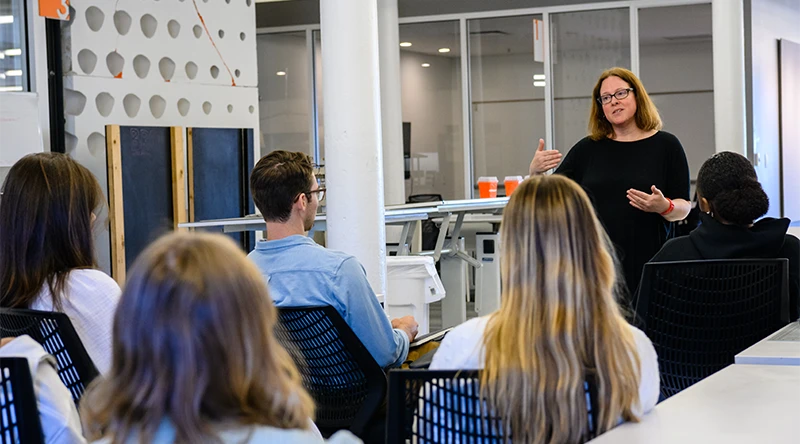A female professor instructs a group of five Clemson students that face her in a room with lots of glass windows.
