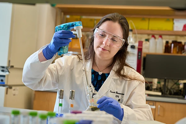 A female graduate student in a lab coat and goggles uses a pipette to fill a bottle.