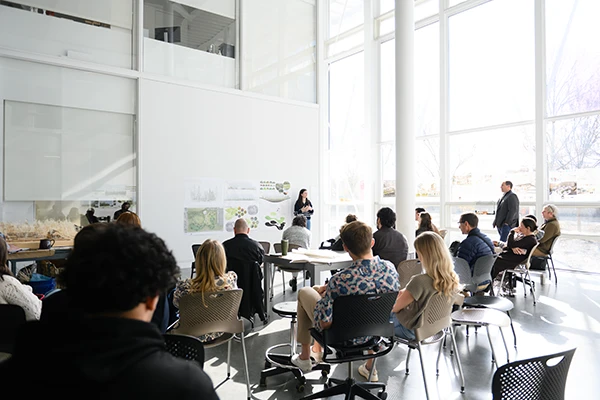 A professor stands in a large white classroom with a wall of glass windows and presents information from a whiteboard to a large group of students.
