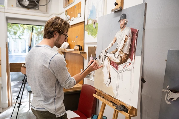 A male Clemson graduate student in an art studio paints a portrait balanced on an easel.