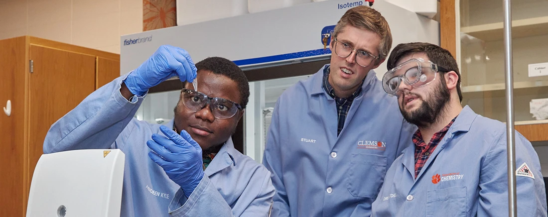 Three male Clemson graduate students in a science lab look at a small container in the leftmost student's hand.
