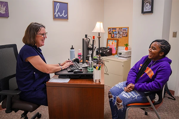 A female student in a Clemson hoodie speaks to a woman that types at a computer behind a desk.