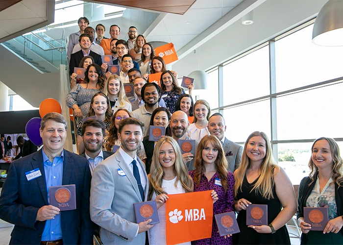 A large group of Clemson MBA students pose with flags and plaques on a staircase.