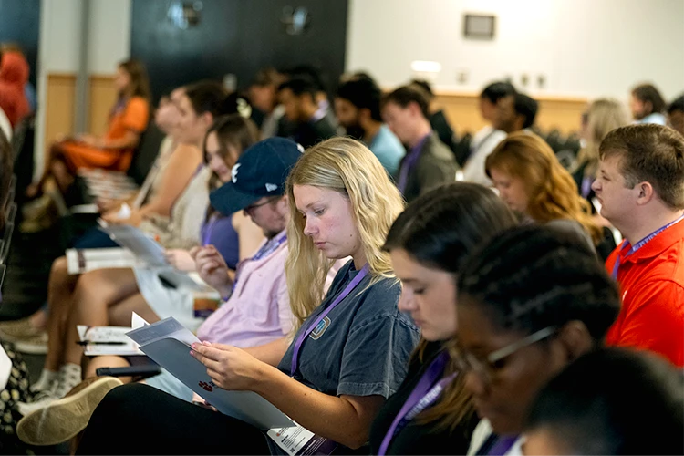 Graduate students sit in rows listening to orientation presentations with printed materials in their laps