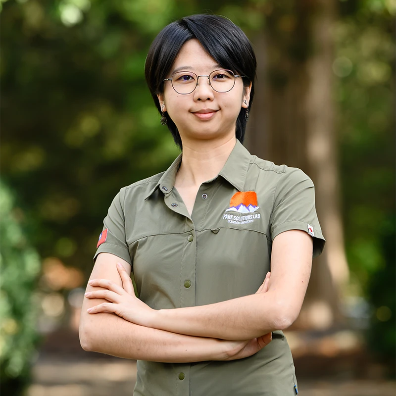 A female PhD student wearing an olive green polo stands with her arms folded with trees in the background.
