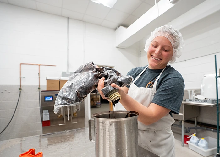 A person in an apron and hair net squeezes a bag of dark liquid into a pot in a kitchen.