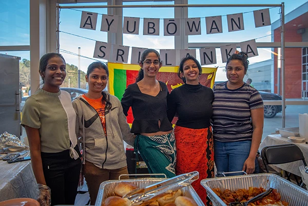 A group of five students pose together in front of a buffet selection of international food, with a banner across the window in the background.