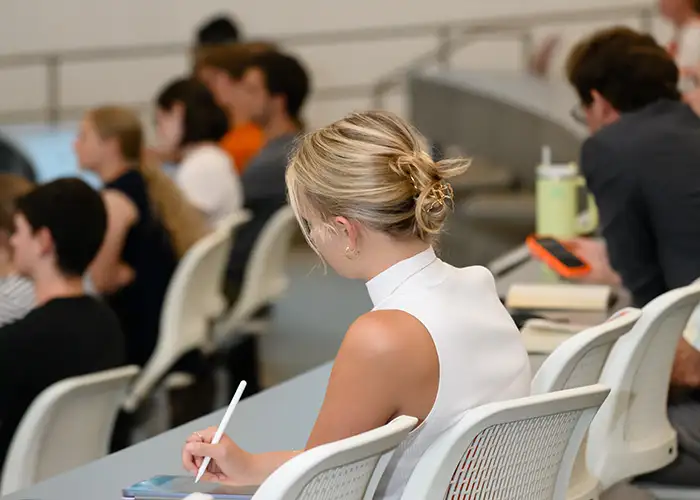 A student takes notes on her tablet during a lecture in a crowded auditorium.