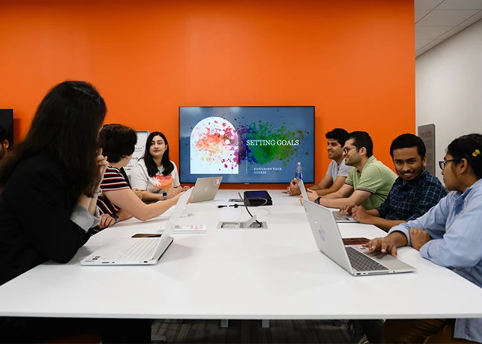 A group of students seated at a long white table with laptops speak to each other, with an orange wall and large TV behind them.