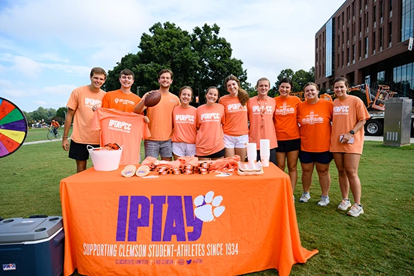 A group of 10 Clemson students in matching orange IPTAY shirts pose together outside.
