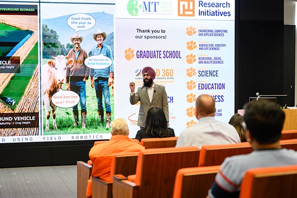A man with a beard presents to an audience while standing in front of a large digital screen.