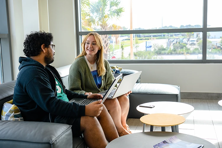 two graduate students talk while sitting on a couch with their laptops