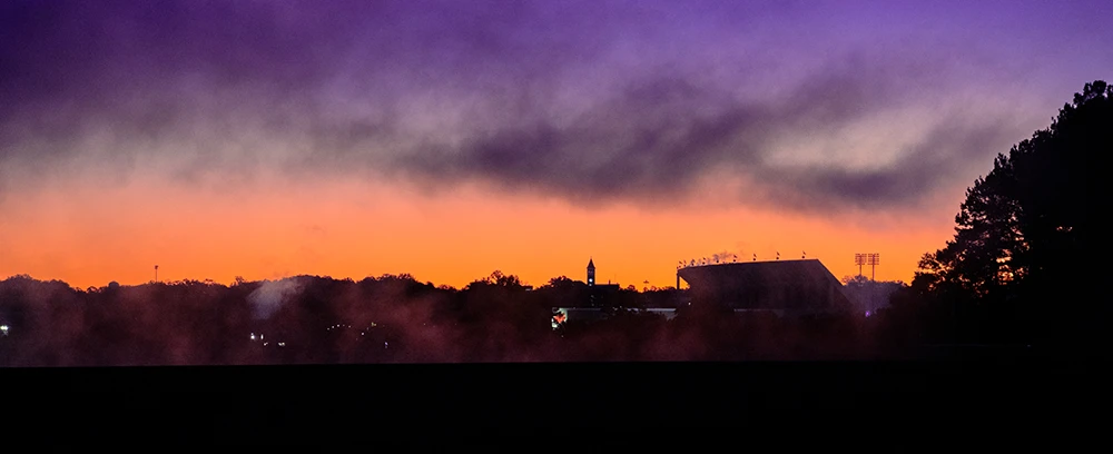 A purple and orange sunset with a tree line and Memorial Stadium in the background.
