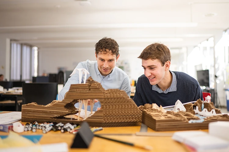 A male professor and male student sit at a table in a studio space with two architectural models.
