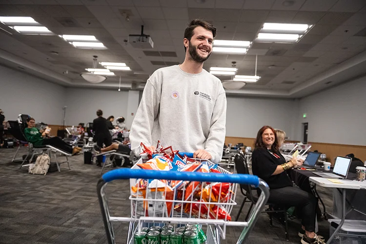 A male student wearing a light gray sweatshirt stands in a computer lab while pushing a grocery cart full of chips and soft drinks.