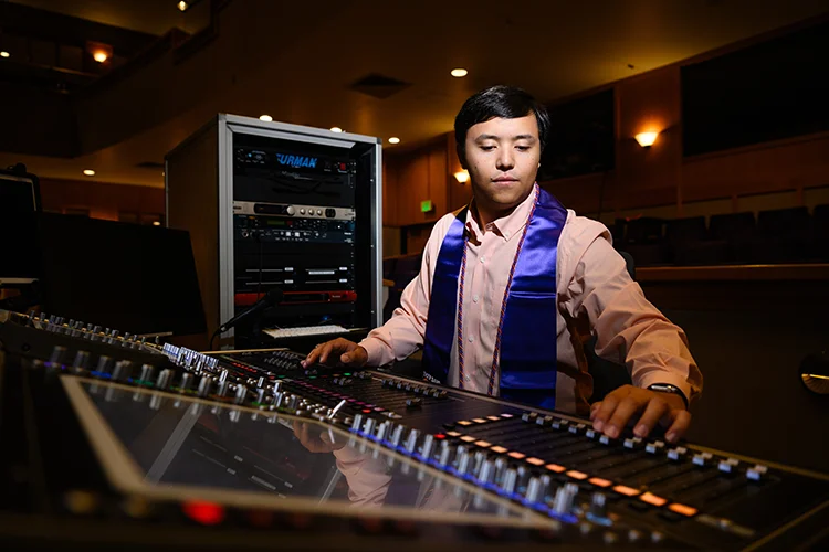 A male student wearing a light orange button-down shirt and purple graduation stole stands in front of an audio switchboard.