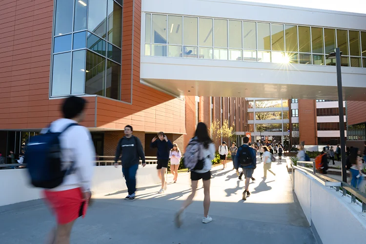 Blurred students walk across a concrete walkway with a brick and glass academic building in the background.