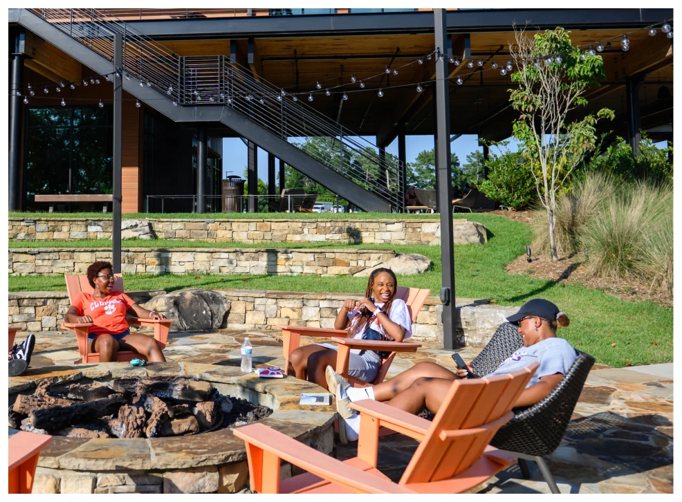Three students sit outdoors on orange Adirondack chairs around a brick fire pit structure.