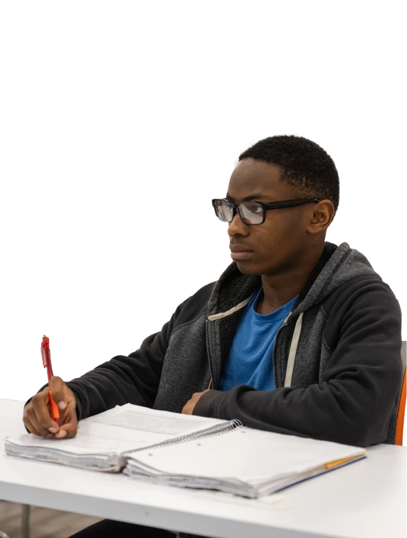 A male student wearing a hooded jacket, T-shirt and glasses sits down and holds a pencil with an open notebook in front of him.
