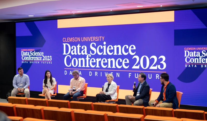 A group of six panelists sits at the front of a room in front of a large screen that says 'Clemson University Data Science Conference 2023: Data-Driven Fun.'
