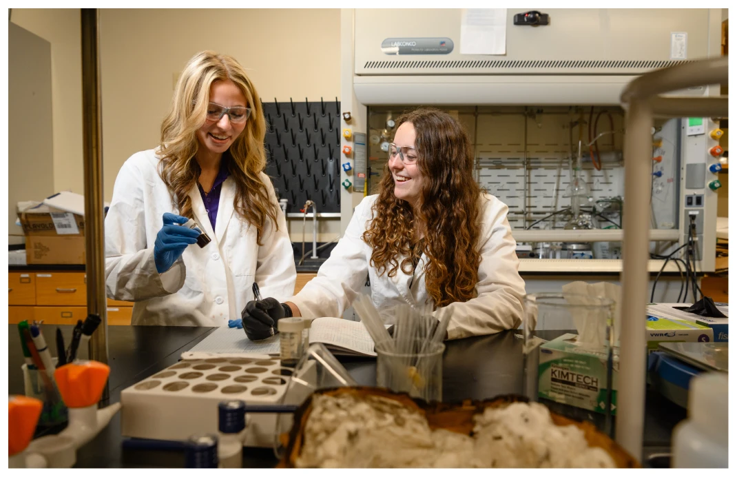 Two women wearing personal protective equipment stand at a table in a laboratory setting and take notes.