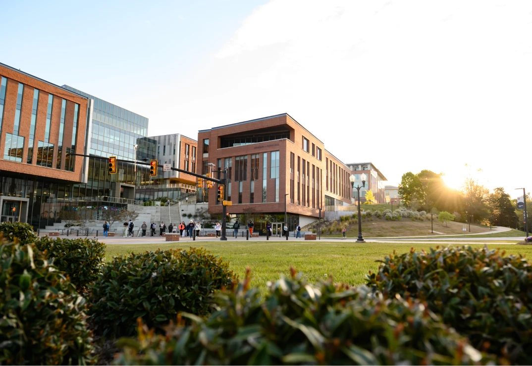 Shrubs in the foreground on the green space of Bowman Field with a brick-and-glass academic building across the street in the background.