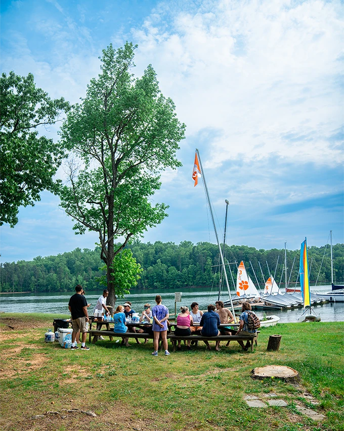 A group of student sits at a picnic table on green grass with a lake and boats in the background on a sunny day.