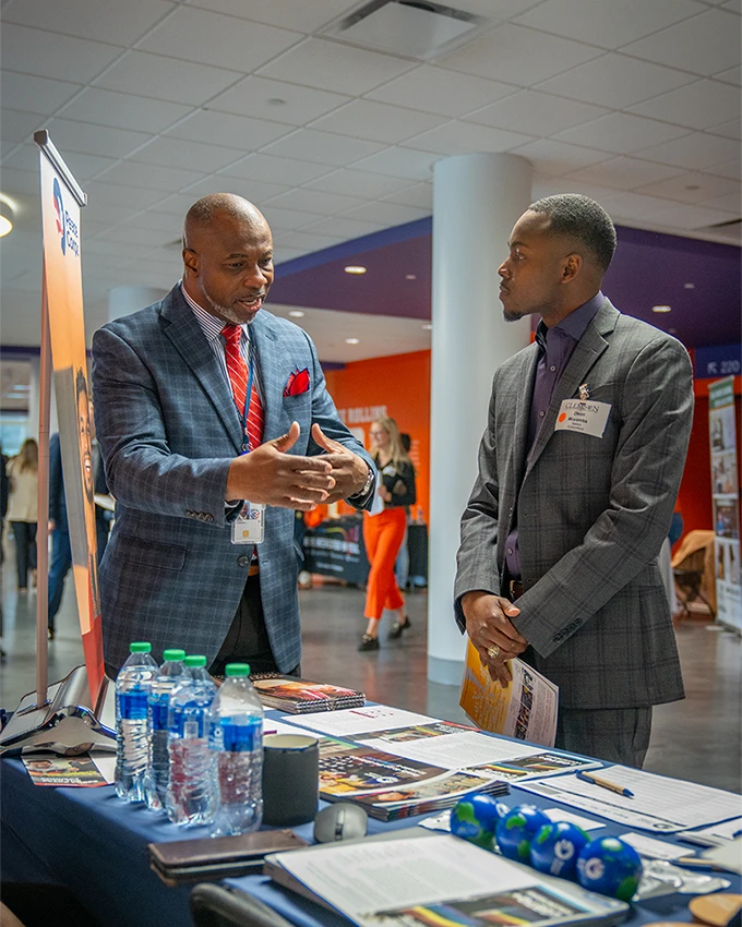 A male student wearing a suit and a male recruiter sit and talk in front of a table of informational pamphlets and brochures during a career fair.