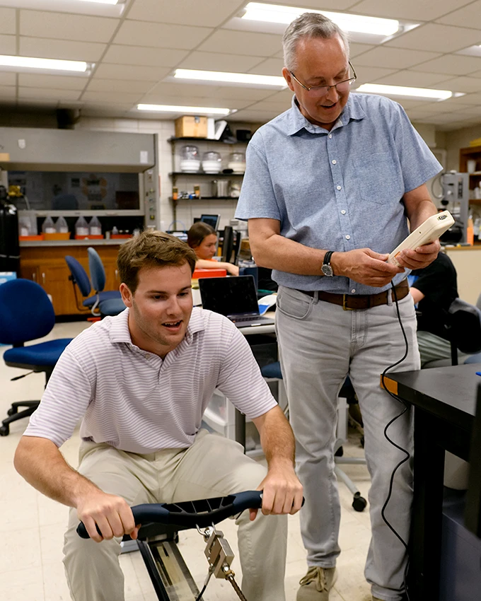 A male student sits at a biking electronic simulation while a male professor operates a corded remote controller.