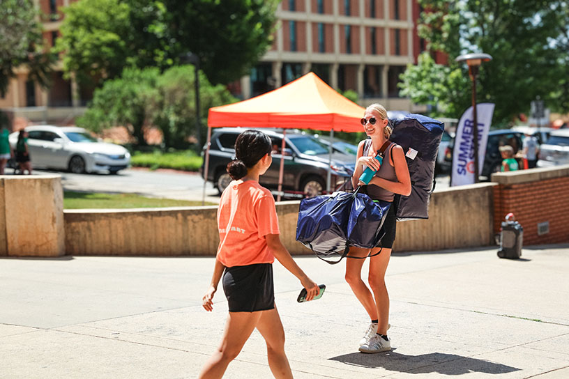 Two female students greet each other in passing while walking on a sidewalk on campus.
