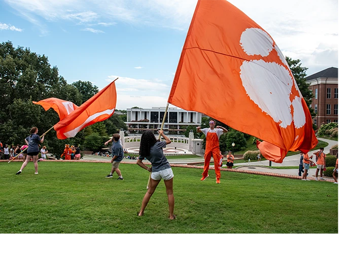 Two students wave large orange Clemson spirit flags on a green lawn with a reflection pond and library in the background.