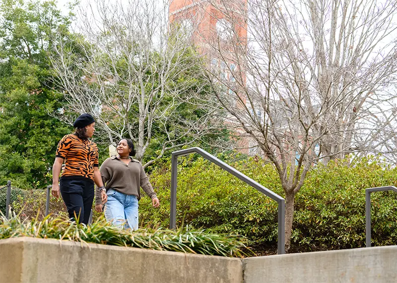 A female student wearing jeans and a brown long-sleeved sweater and a female staff member wearing black pants and a tiger-stripe short-sleeved sweater and a black hat have a conversation on a campus sidewalk.