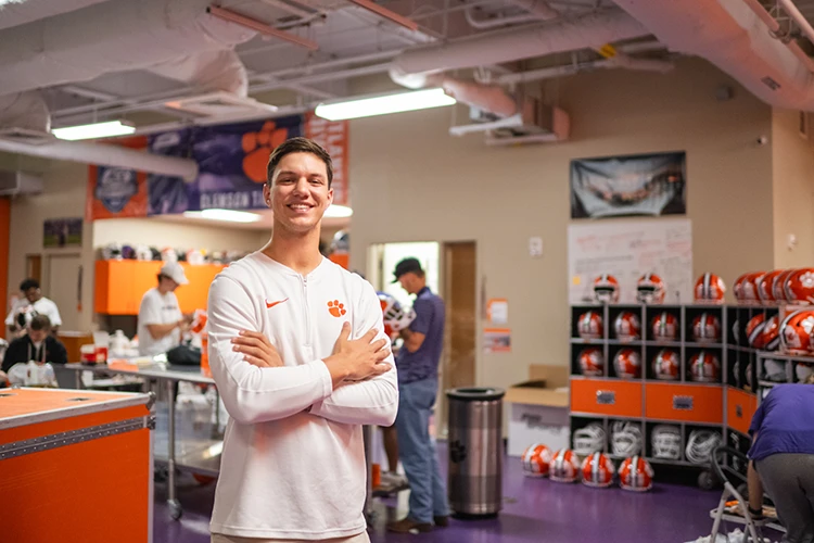 A male student wearing a white long-sleeved T-shirt with an orange Clemson Paw and Nike swoosh logos stands with his arms folded in a football equipment room space.