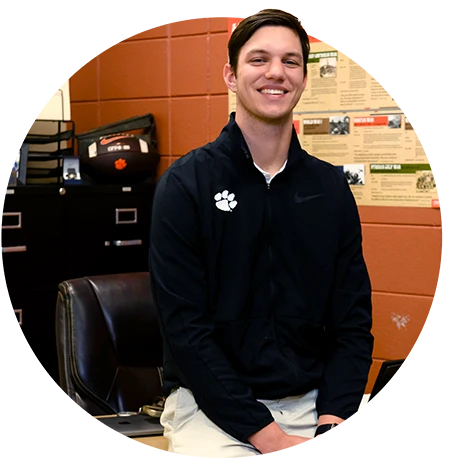 A male student teacher wearing a black quarter-zip sweater with a white Clemson Tiger Paw logo sits on a desk and smiles in a high school classroom.