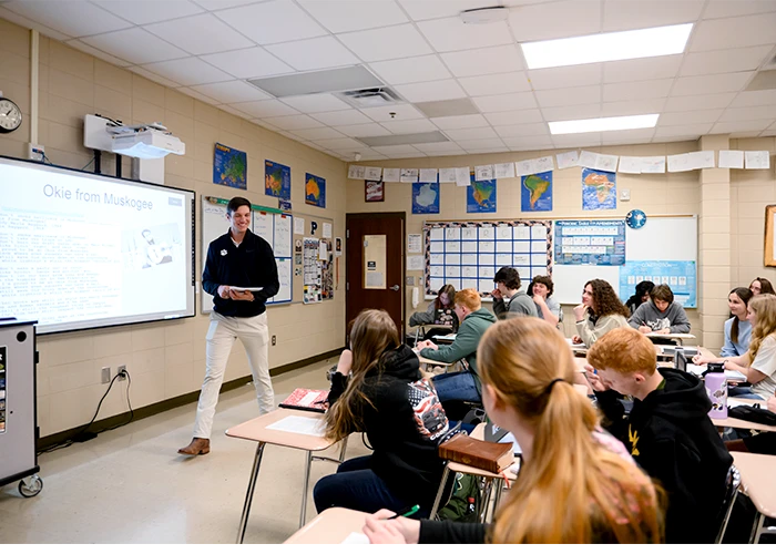 A male student teacher wearing khaki pants and a black quarter-zip sweater with a white Clemson Tiger Paw logo stands at the front of a high school classroom and instructs students.